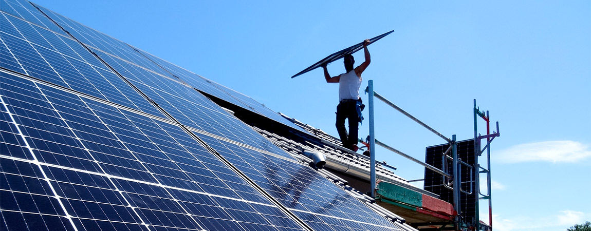 Hamacher Arbeitskraft steht mit einer Solarzelle über dem Kopf auf einem Gerüst vor einem mit Solarzellen bedeckten Dach
