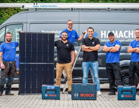 Vier Männer in Hamacher Kleidung und mit einer Solarzelle stehen vor einem Van mit Hamacher Aufdruck und lächeln in die Kamera